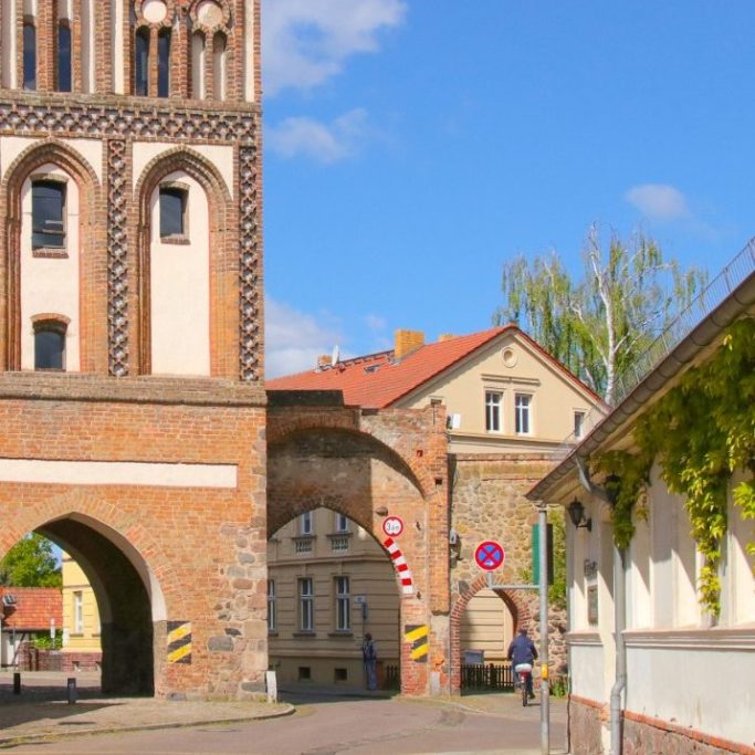 Alter Tor mit Ziegelmauer, umgeben von architektonischen Gebäuden und klarem Himmel. Durch das Tor ist die Logopädische Praxis erkennbar.