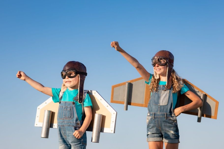 Zwei Kinder mit selbstgebauten Flügeln und Fliegerhelmen, die mutig in den Himmel blicken.