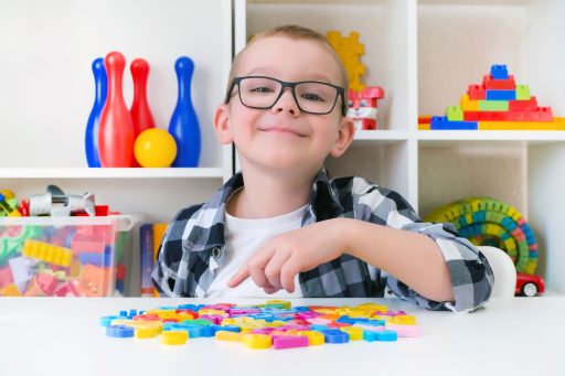 Ein Junge mit Brille lächelt und zeigt auf bunte Spielsteine auf einem Tisch.